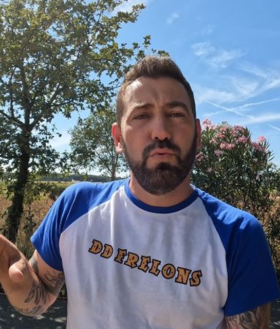 Portrait extérieur d'un homme barbu et tatoué, portant un t-shirt DD FRELONS (bleu et blanc) devant la végétation et un ciel d'été.